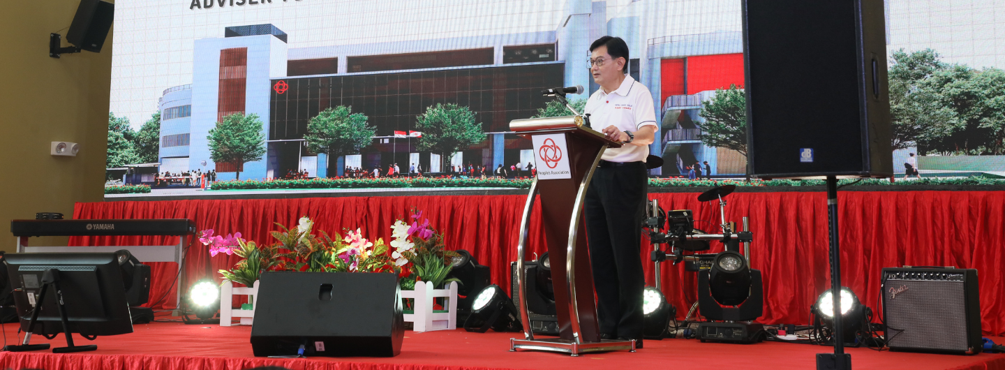 Man at lectern with "People's Association" logo, building image backdrop, stage lights, Yamaha keyboard, Fender amp.